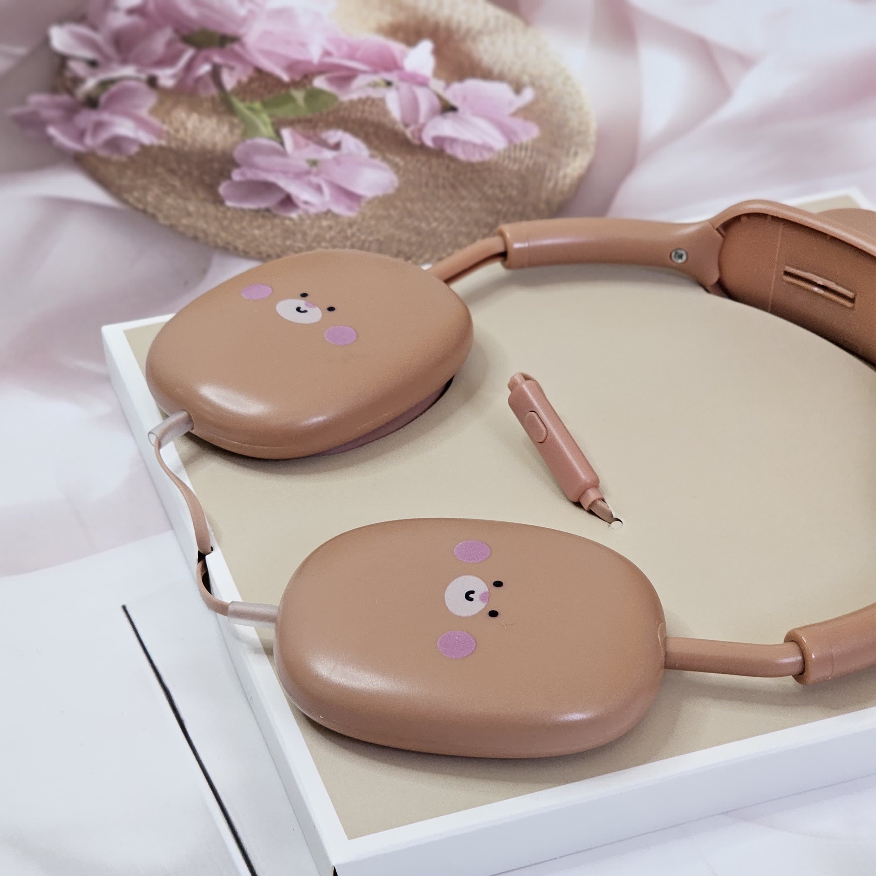 Close-up of pink BuBu Bear wired headphones for kids, showcasing the rabbit-inspired design with bear face on the earcups, a detachable microphone, and the headphones positioned on a white surface with floral accents. www.usmile.toys